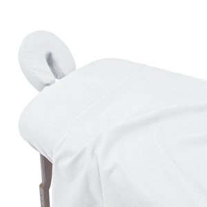 May include: White massage table with a white face cradle. The table is covered in a white sheet.
