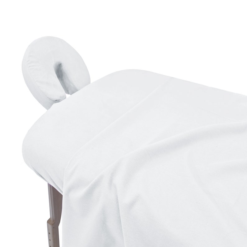 May include: White massage table with a white face cradle. The table is covered in a white sheet.