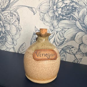 May include: A round, speckled beige ceramic vinegar cruet with a cork stopper. The vessel features a textured, light brown rectangular label with the word Vinegar embossed in dark lettering. It has a narrow neck and a small handle on the side.