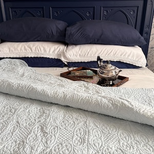 May include: A light blue quilted bedspread with an intricate stitched pattern covers a bed featuring navy blue and white pillows with lace trim. A dark wooden tray sits on the bed, holding a silver teapot and a small book with a colourful cover.
