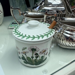May include: A white ceramic jam pot with a lid, featuring a green leaf border pattern on the lid and a botanical illustration of daisies and the text Bellis Perennis on the side. A small wooden spoon is inserted through a notch in the lid.