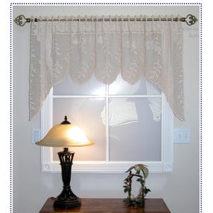 May include: A cream-colored crochet valance featuring a wheat pattern, hanging from a decorative bronze-toned curtain rod. The valance has a scalloped bottom edge and is displayed over a window. The text "Blowing Wheat Valance" is visible.