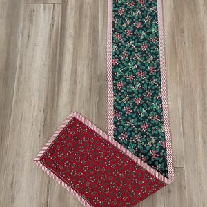 May include: A Christmas-themed table runner with a red and green color scheme. The runner features a dark green fabric with holly and candy cane patterns, and a red fabric with a wreath pattern. Both are bordered with red and white stripes.