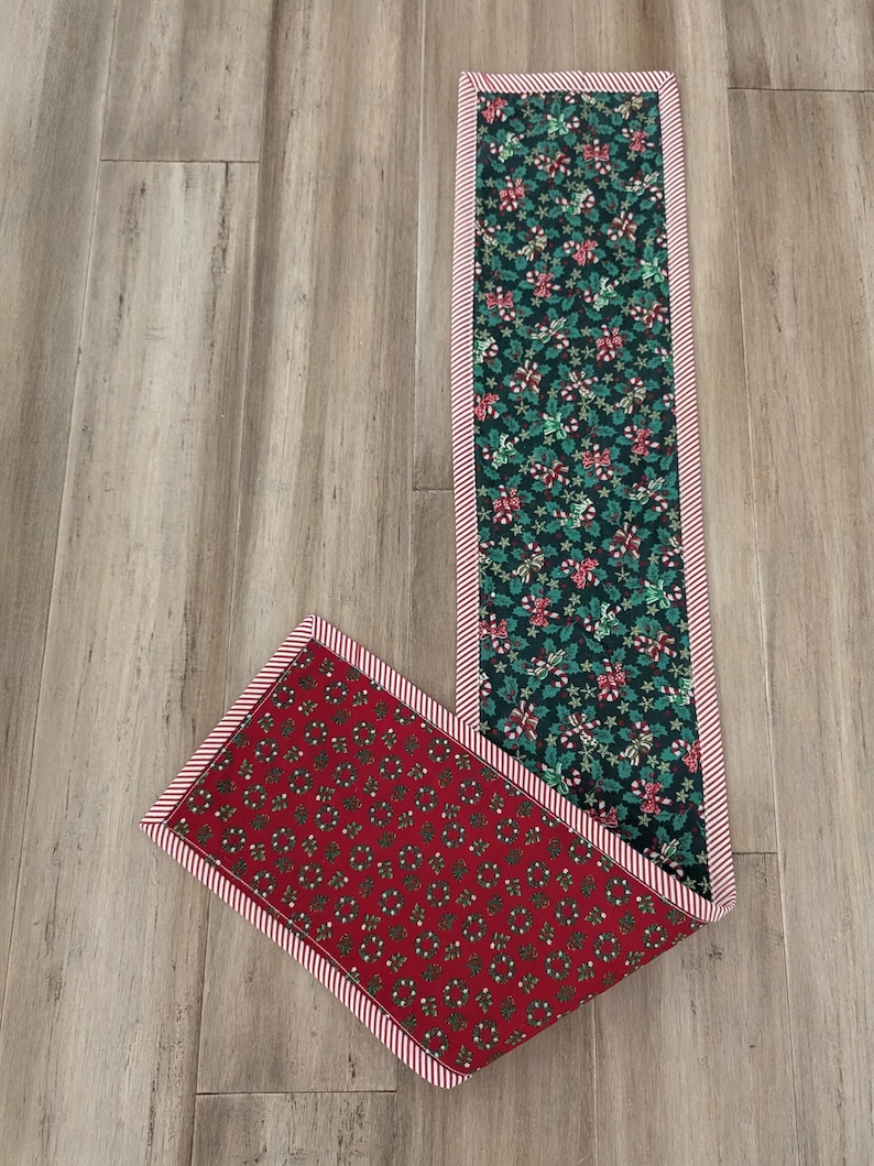 May include: A Christmas-themed table runner with a red and green color scheme. The runner features a dark green fabric with holly and candy cane patterns, and a red fabric with a wreath pattern. Both are bordered with red and white stripes.