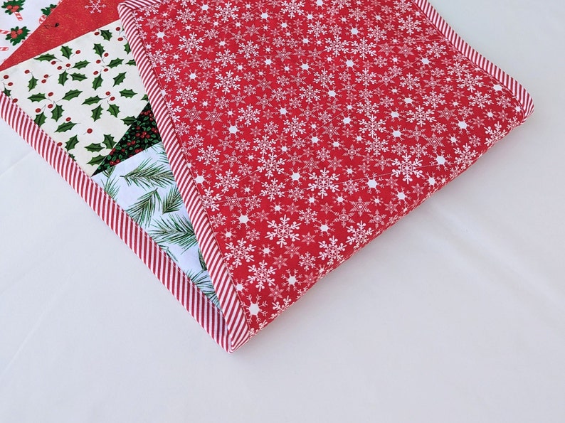 May include: A Christmas-themed quilted placemat with a red snowflake pattern and a striped border. Other sections feature holly, pine needles, and a red and white candy cane design. The placemat is square.