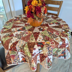May include: A round, patchwork tablecloth with a fall theme. The fabric features triangles of various patterns, including leaves, pumpkins, and floral designs in shades of brown, orange, and green. A pumpkin holds a floral arrangement.