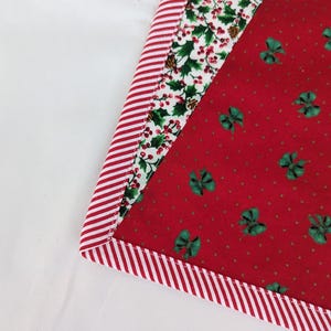 May include: A Christmas-themed fabric item with a red and white striped border. The item features a red background with green bow accents and a white background with holly and berry patterns. The item is likely a placemat or a similar decorative piece.