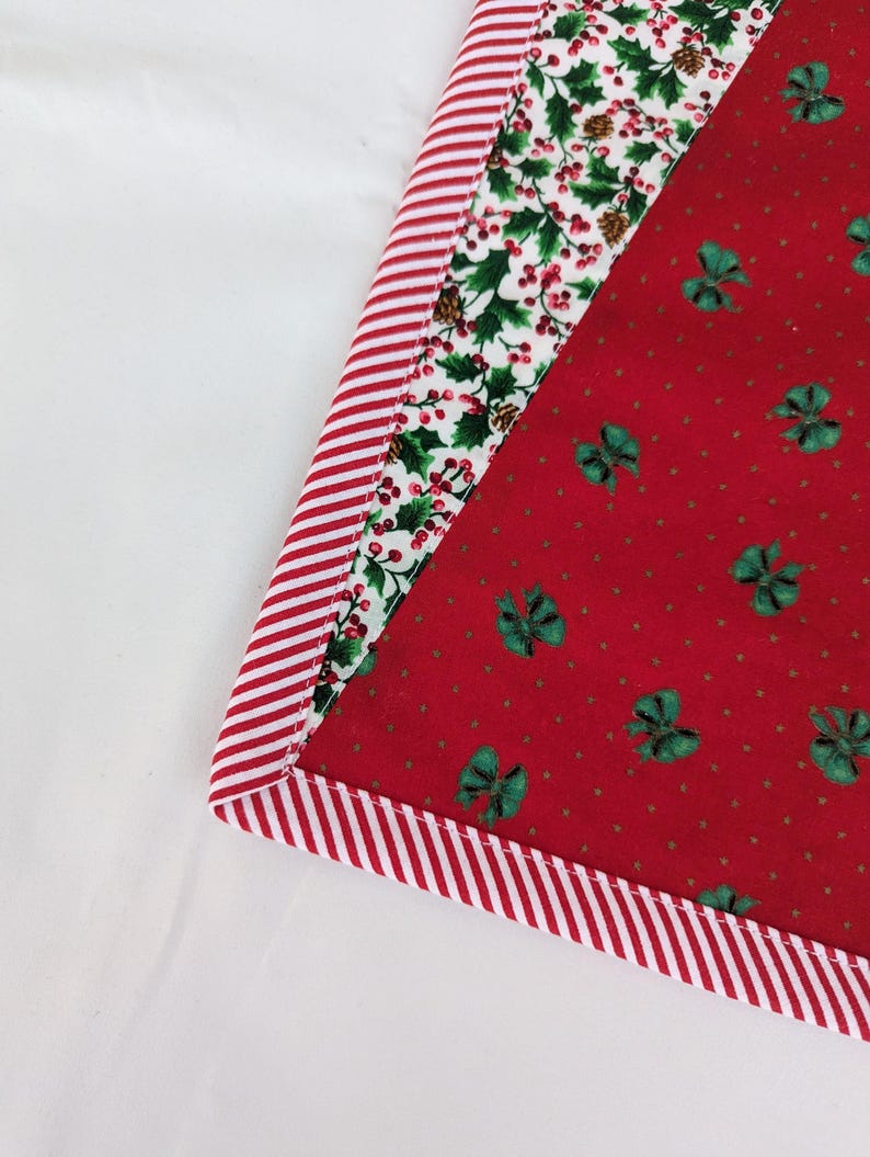May include: A Christmas-themed fabric item with a red and white striped border. The item features a red background with green bow accents and a white background with holly and berry patterns. The item is likely a placemat or a similar decorative piece.