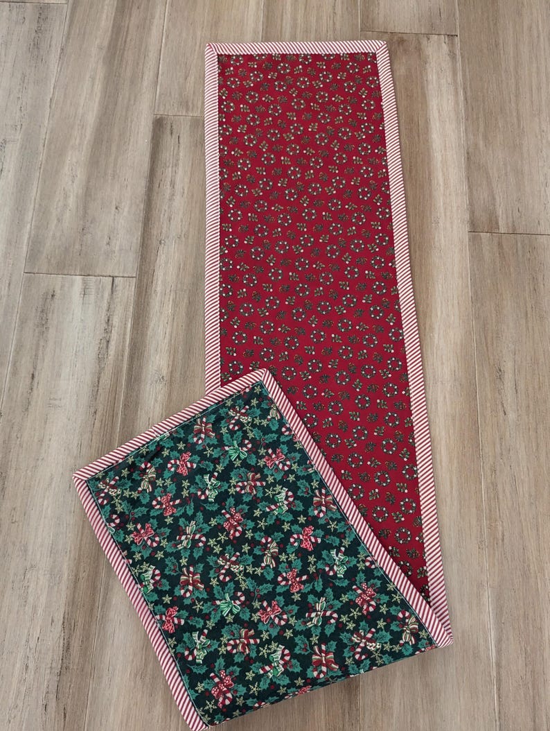 May include: Two Christmas-themed quilted table runners. One is rectangular with a red background and a pattern of small white and green floral designs. The other is square with a dark green background and a pattern of candy canes, holly, and stars. Both have a red and white striped border.