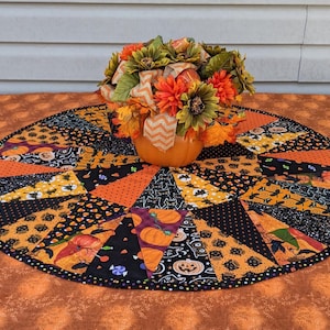 May include: A round, quilted Halloween table topper with a pumpkin centerpiece. The topper features orange, black, and yellow fabric with Halloween-themed patterns. The pumpkin holds orange and green flowers with a decorative ribbon.