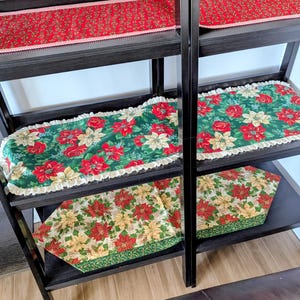 May include: A black shelving unit displays several Christmas table runners. The runners feature festive patterns of red poinsettias, roses, and greenery on green and cream backgrounds. Some runners have a ruffled edge.