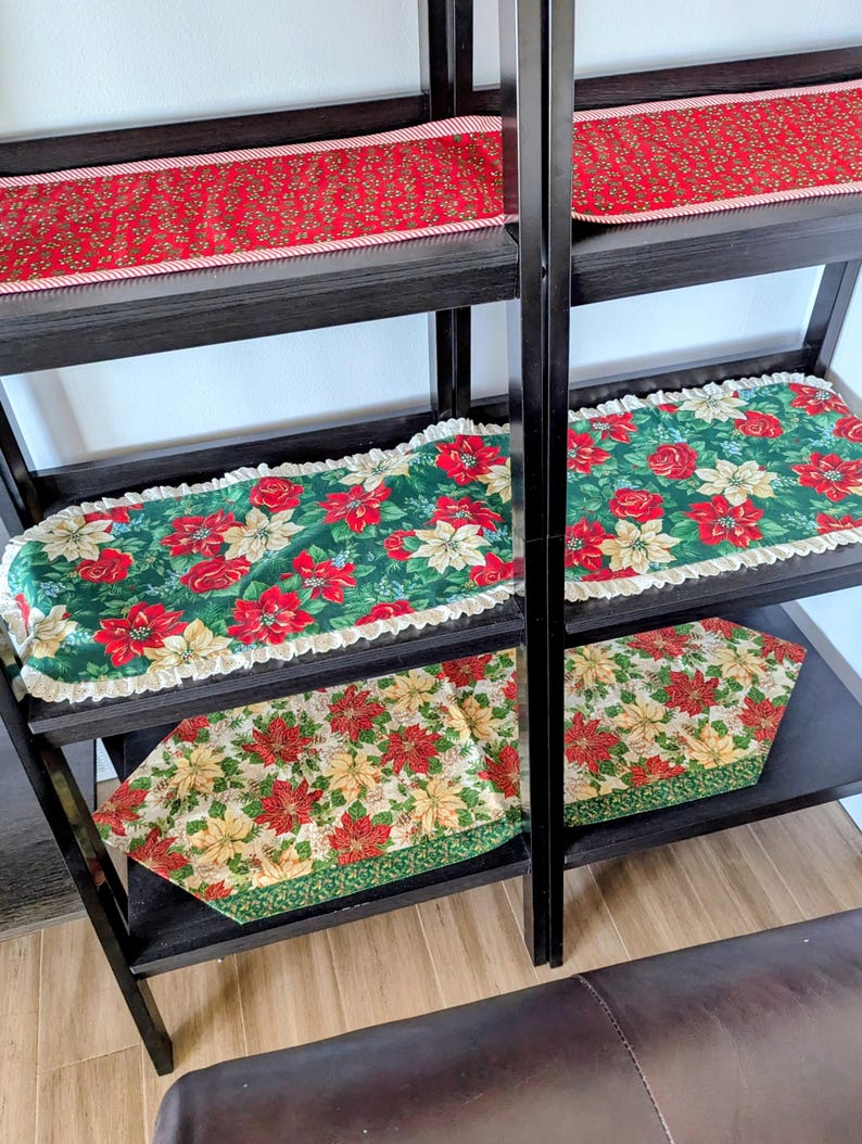 May include: A black shelving unit displays several Christmas table runners. The runners feature festive patterns of red poinsettias, roses, and greenery on green and cream backgrounds. Some runners have a ruffled edge.