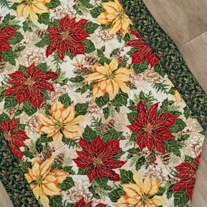 May include: A Christmas-themed table runner featuring a pattern of red and yellow poinsettias, green leaves, and pine cones on a cream background. The runner has a dark green border with a holly and berry design. A festive holiday decoration.