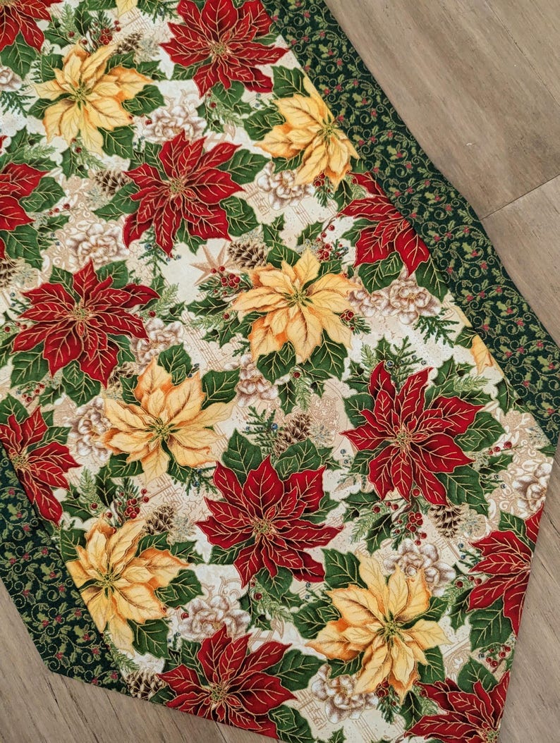 May include: A Christmas-themed table runner featuring a pattern of red and yellow poinsettias, green leaves, and pine cones on a cream background. The runner has a dark green border with a holly and berry design. A festive holiday decoration.