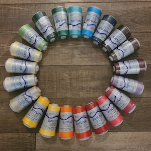 May include: A circular arrangement of colorful spools of SureLock thread. The spools are in various shades, including green, blue, yellow, orange, and red. The spools are arranged on a wooden surface.