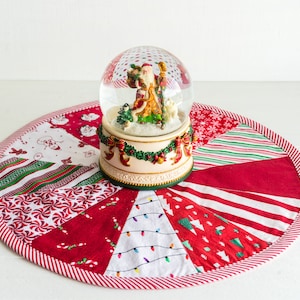 May include: A red and white patchwork Christmas tree skirt with a snow globe featuring Santa Claus in the center.