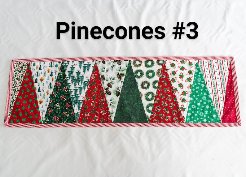 May include: A Christmas-themed table runner with a variety of festive patterns. The runner features alternating triangular sections with designs like Christmas trees, wreaths, and candy canes. The border is red and white striped, and the text "Pinecones #3" is at the top.