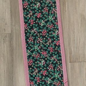 May include: A rectangular Christmas table runner with a dark green background featuring a pattern of red and green bows, holly leaves, and stars. The edges are trimmed with a red and white striped border. The runner is approximately 12 inches wide and 48 inches long.