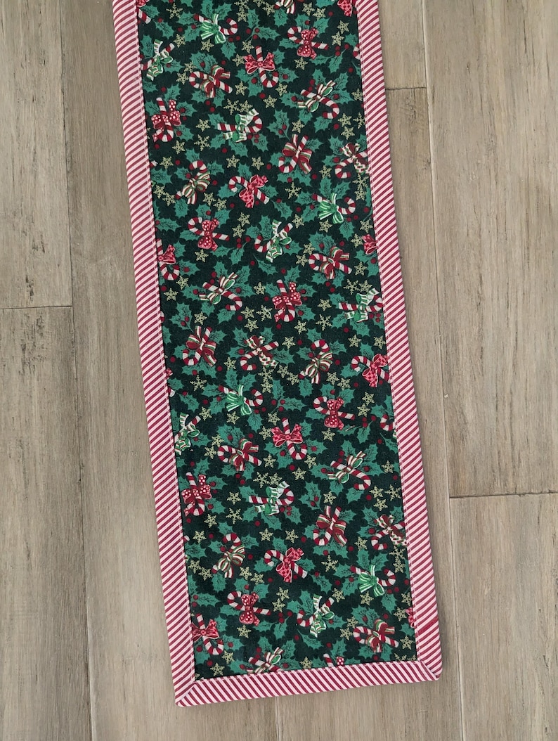May include: A rectangular Christmas table runner with a dark green background featuring a pattern of red and green bows, holly leaves, and stars. The edges are trimmed with a red and white striped border. The runner is approximately 12 inches wide and 48 inches long.