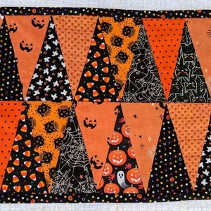 May include: A rectangular Halloween-themed placemat with a patchwork design. The triangles feature orange, black, and white fabrics with patterns of pumpkins, cats, candy corn, and polka dots. The border is black with small, colorful polka dots.