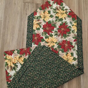 May include: A Christmas-themed table runner with a poinsettia floral pattern. The runner features red and yellow poinsettias on a cream background, with a green border. The reverse side has a green holly leaf pattern.