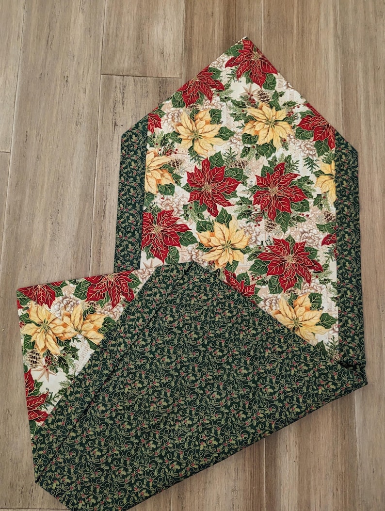 May include: A Christmas-themed table runner with a poinsettia floral pattern. The runner features red and yellow poinsettias on a cream background, with a green border. The reverse side has a green holly leaf pattern.