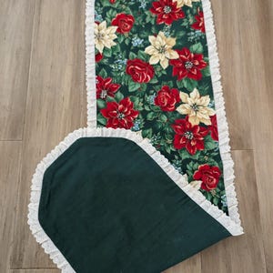 May include: A Christmas-themed table runner with a dark green background and a floral pattern of red poinsettias, roses, and cream-colored flowers. The runner is trimmed with white lace.
