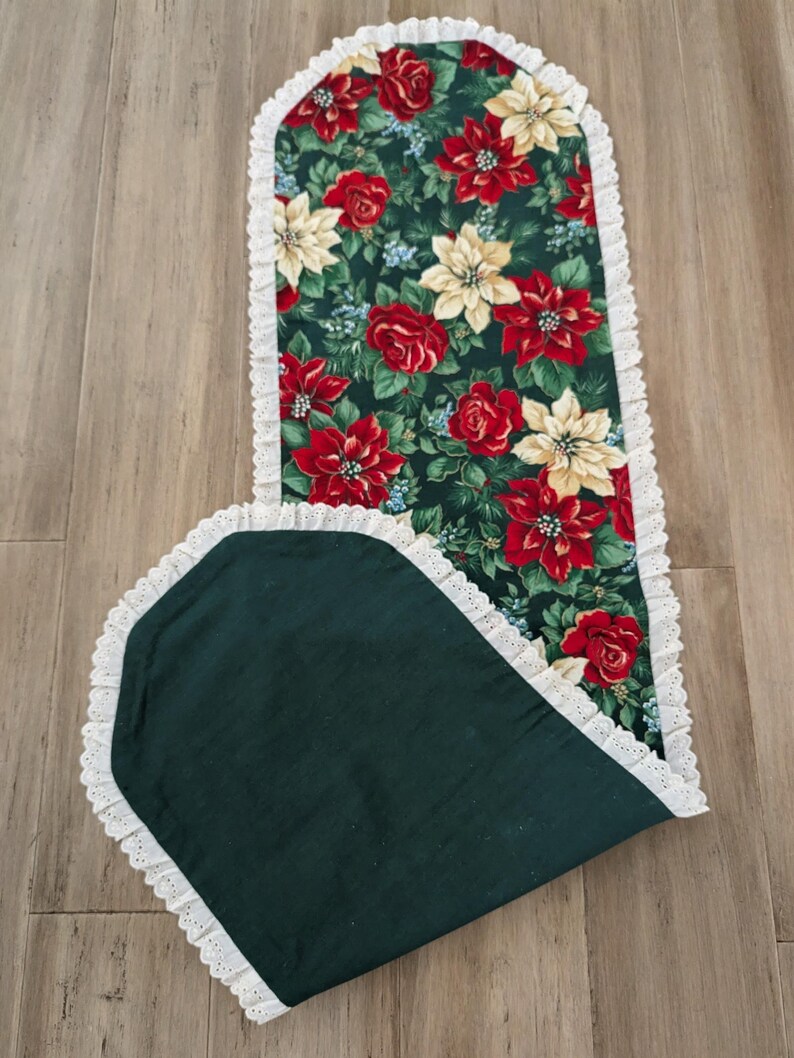 May include: A Christmas-themed table runner with a dark green background and a floral pattern of red poinsettias, roses, and cream-colored flowers. The runner is trimmed with white lace.