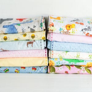 May include: A stack of folded fabric swaddles featuring various colorful patterns including dinosaurs, bees, horses, trains, zoo animals, frogs, crocodiles, and strawberries. The textiles display diverse prints on light backgrounds with stitched edges.