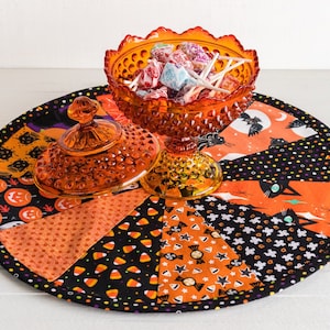 May include: An orange glass candy dish filled with lollipops sits on a round, quilted Halloween-themed placemat. The placemat features orange, black, and white patterned fabric with Halloween motifs like pumpkins, bats, and candy corn.