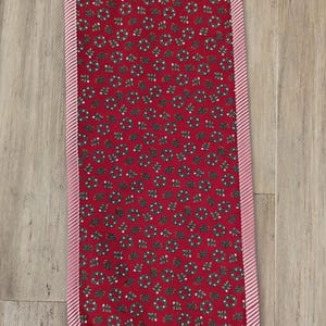 May include: A rectangular red table runner with a repeating pattern of green wreaths and white berries. The runner is trimmed with a pink and white striped border, creating a festive holiday aesthetic.