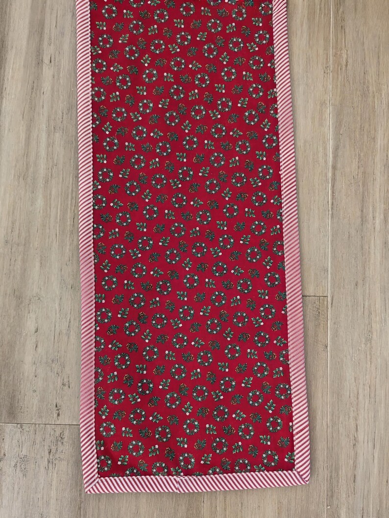May include: A rectangular red table runner with a repeating pattern of green wreaths and white berries. The runner is trimmed with a pink and white striped border, creating a festive holiday aesthetic.