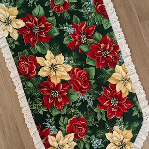 May include: A festive table runner featuring a dark green background adorned with red poinsettias, yellow flowers, and red roses. The edges are trimmed with a white lace border, creating a decorative holiday accent.