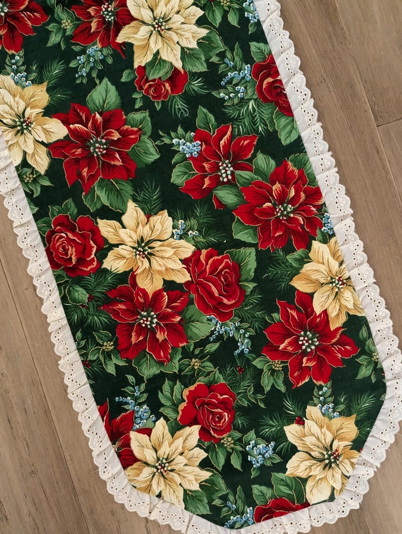 May include: A festive table runner featuring a dark green background adorned with red poinsettias, yellow flowers, and red roses. The edges are trimmed with a white lace border, creating a decorative holiday accent.