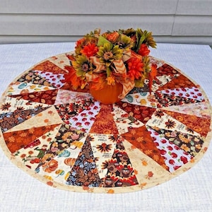 May include: A round, quilted table topper with a patchwork design in shades of orange, brown, and cream. The topper features various fall-themed patterns, including pumpkins, leaves, and turkeys. A small pumpkin holds a floral arrangement.