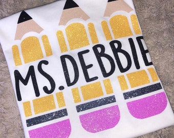 May include: White t-shirt with three yellow, black, and pink glitter pencils and the text "MS. DEBBIE" in black.