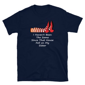 Puede incluir: Camiseta azul marino con un gráfico blanco de un par de piernas rayadas rojas y blancas con zapatos rojos. El texto "I Haven't Been The Same Since That House Fell on My Sister" está impreso debajo del gráfico.