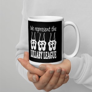 May include: White ceramic mug with a black background and white text that reads "We represent the LULLABY LEAGUE". There are three white ballet shoes hanging upside down above the text.