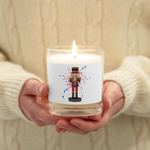 May include: A white candle with a nutcracker design on the label. The nutcracker is holding a drum and is surrounded by colorful confetti. The candle is lit and the flame is visible.