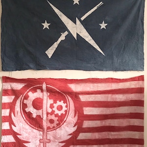 Op de afbeelding: Twee vlaggen hangen aan een muur. De bovenste vlag is blauw met een witte bliksem, twee sterren en een gekruist geweer en schroevendraaier. De onderste vlag is rood met witte strepen en een wit symbool van een zwaard en tandwielen.