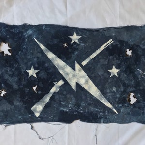 Könnte beinhalten: Eine verblasste blaue Flagge mit einem weißen Blitz und gekreuzten Gewehren. Auf der Flagge sind fünf weiße Sterne zu sehen.