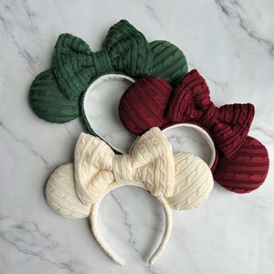 May include: Three headbands featuring round ears and a large central bow, all covered in a cable knit fabric. The headbands are available in dark green, deep burgundy, and cream colors, each with a white inner lining on the headband frame.