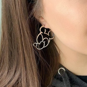 May include: Silver-coloured Mickey Mouse-shaped earrings. The earrings are a cutout design, showcasing the iconic character's head and face. The earrings are worn on the ear, with the post visible.