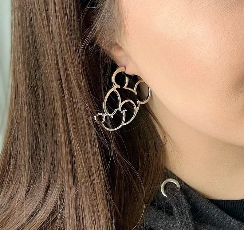 May include: Silver-coloured Mickey Mouse-shaped earrings. The earrings are a cutout design, showcasing the iconic character's head and face. The earrings are worn on the ear, with the post visible.