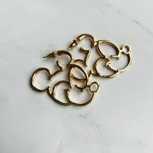 May include: A pair of gold-coloured hoop earrings shaped like Mickey Mouse. The earrings are made of shiny metal and rest on a white and grey marble surface. They have a post and back closure.