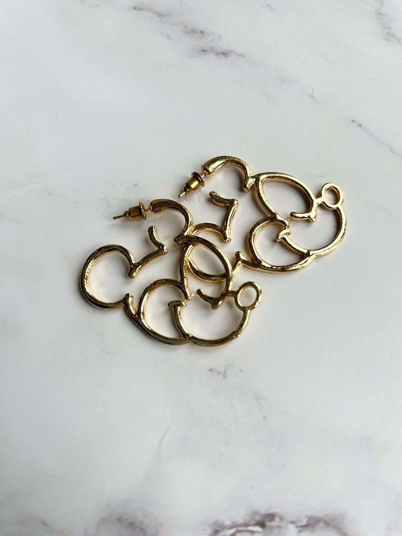 May include: A pair of gold-coloured hoop earrings shaped like Mickey Mouse. The earrings are made of shiny metal and rest on a white and grey marble surface. They have a post and back closure.