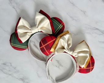 May include: Two headbands featuring round ears with plaid fabric patterns. One headband has green and red plaid ears with a cream satin bow. The second headband has red and yellow plaid ears with a gold-toned satin bow. Both headbands have white bands.