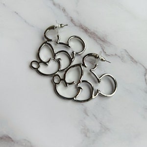 May include: Silver-coloured hoop earrings shaped like Mickey Mouse. The earrings are made of shiny metal and feature the iconic mouse silhouette. The earrings are displayed on a white and grey marble surface.