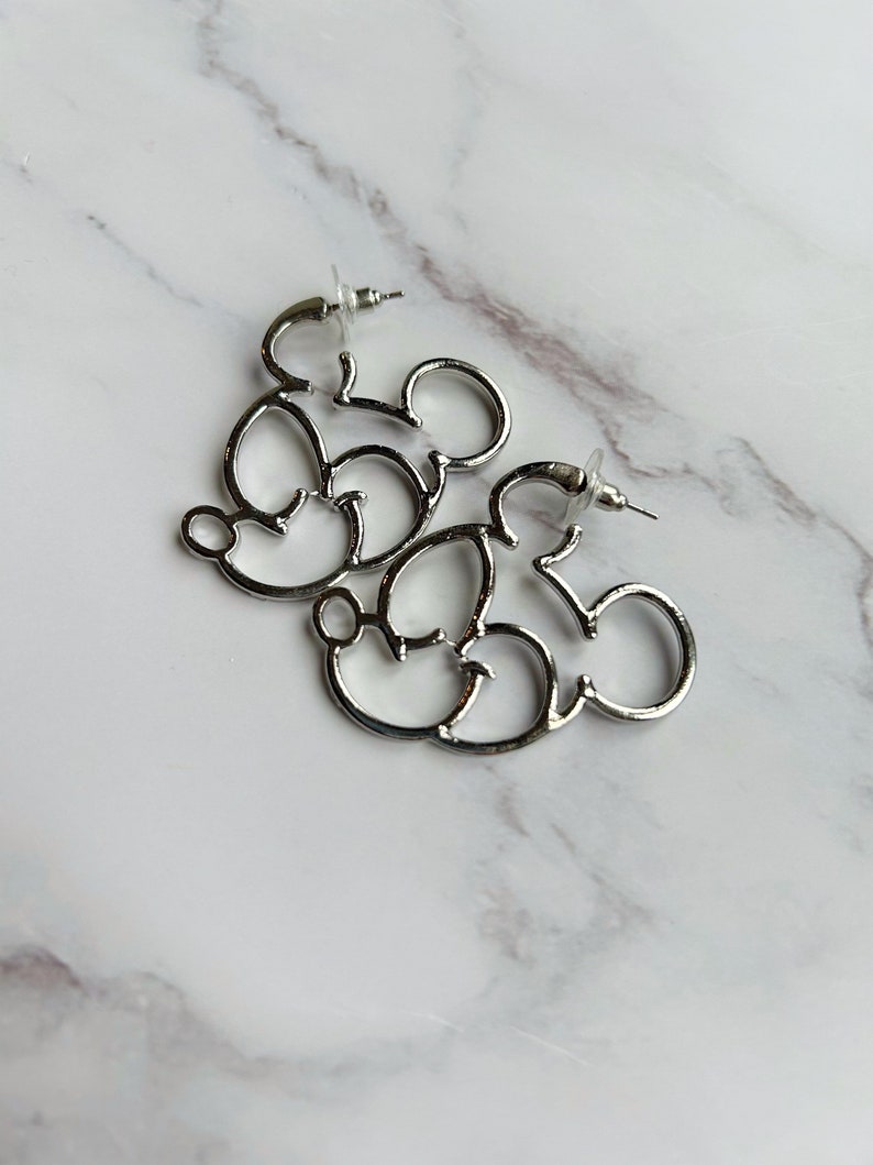May include: Silver-coloured hoop earrings shaped like Mickey Mouse. The earrings are made of shiny metal and feature the iconic mouse silhouette. The earrings are displayed on a white and grey marble surface.