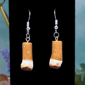 Op de afbeelding: Een paar zilveren oorhangers met een realistische sigaretbedel. De sigaretbedels zijn bruin en wit met een wit filter.
